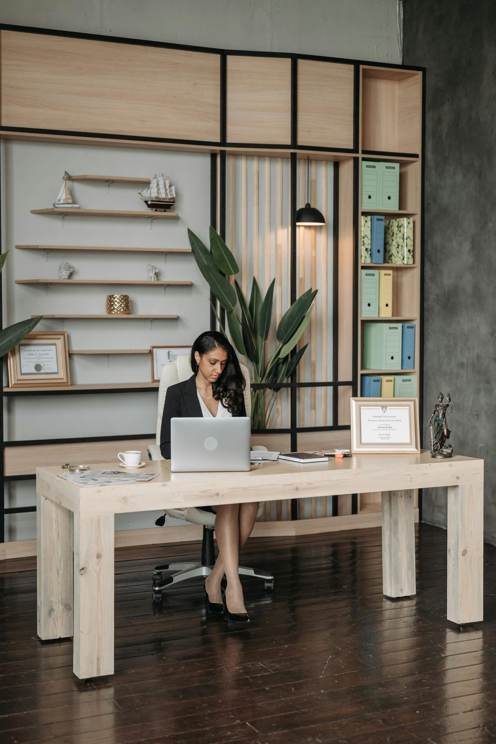 Woman in office working on a laptop at a modern wooden desk with shelves and decor.